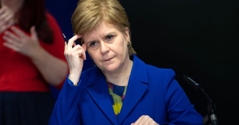 Scotland's first minister Nicola Sturgeon wears a blue and green top with a matching blue jacket during a press conference. Her head is resting against her hand