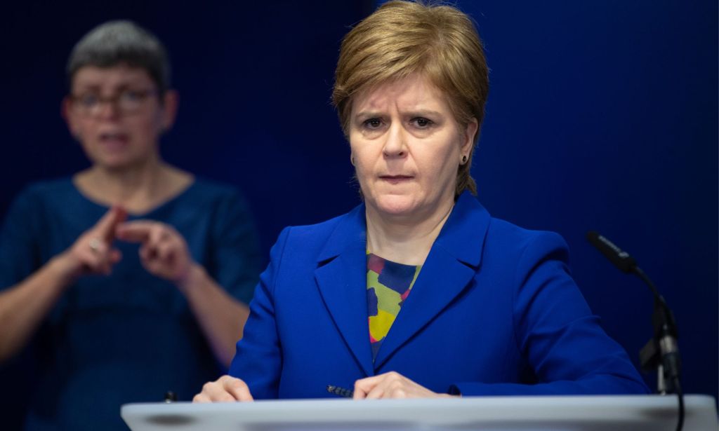 Scotland's first minister Nicola Sturgeon wears a blue and green top with a matching blue jacket during a press conference. She is standing at a podium