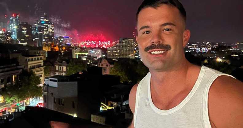 Simon, a white man with a moustache, smiling while stood on a balcony in front of a nighttime skyline