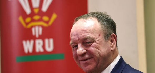 Steve Phillips wears a white button-up shirt and blue jacket as he stands in front of a red banner displaying the Welsh Rugby Union logo and WRU lettering