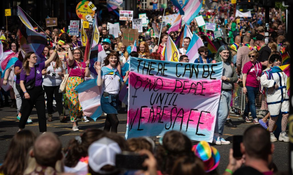 A person in the middle of a trans Pride march holds up a sign reading 'There can be no peace until we are safe' in the colours of the trans Pride flag