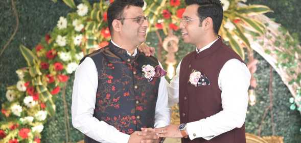 Abhay and Supriyo, two Indian men, looking lovingly at each other in front of a wall of flowers