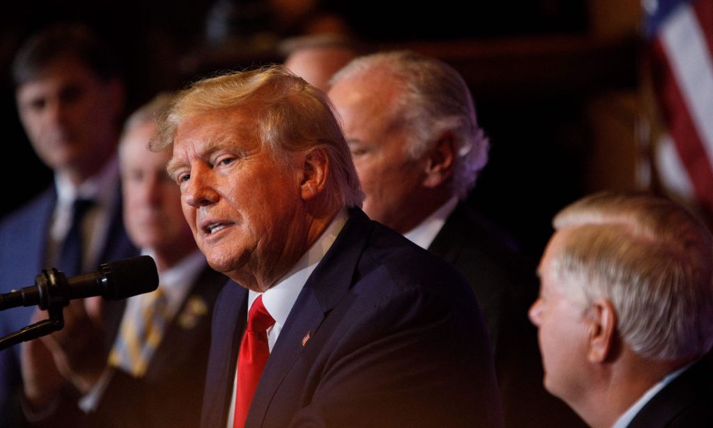 A photo shows Donald Trump wearing a navy suit, whit shirt and red tie speaking to an audience during a campaign speech.