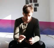 Man sitting on bed looking at phone with bisexual Pride flag colours behind him