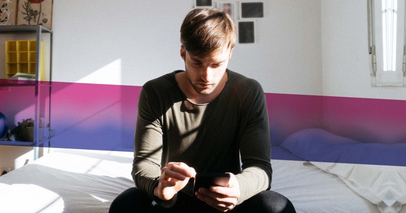 Man sitting on bed looking at phone with bisexual Pride flag colours behind him
