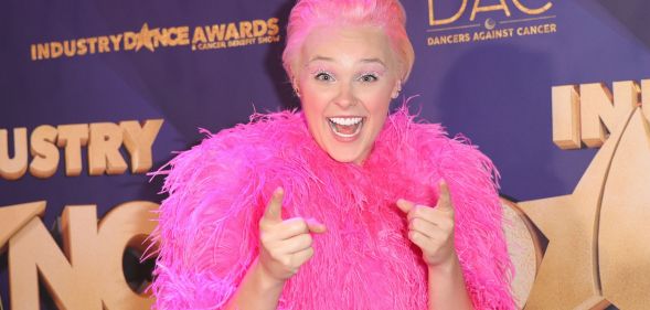 JoJo Siwa on the 2022 Industry Dance Awards red carpet, wearing a pink fluffy jacket and slicked back blonde hair. JoJo is smiling and pointing at the camera.