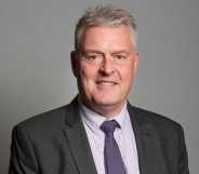 Lee Anderson pictured in his official parliamentary portrait. He is wearing a grey suit with a dark coloured tie and is standing against a grey background.