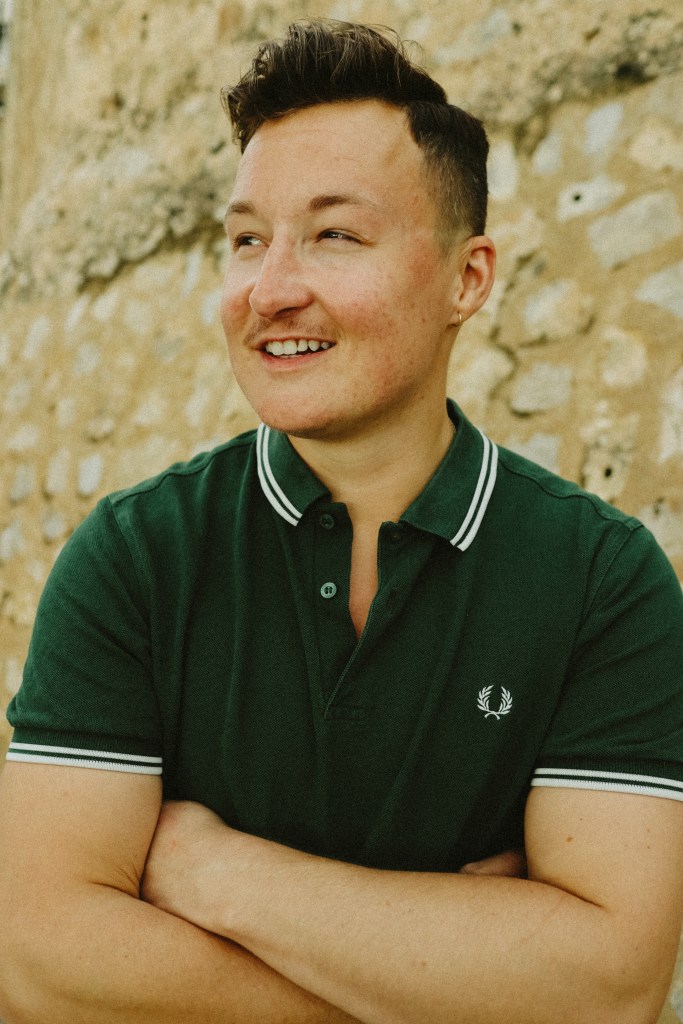 Max Siegel is wearing a green polo shirt, and with their arms folded, they are smiling off to the left of the camera. 