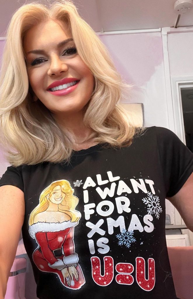 Rebecca Tallon de Havilland wearing a t-shirt with the slogan "All I want for Xmas is U=U". She has long wavy blonde hair and is pictured indoors.