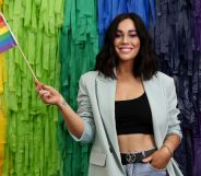 Sophie Cachia wearing a black top and grey blazer stands with one hand in her pocket and the other holding a pride flag.