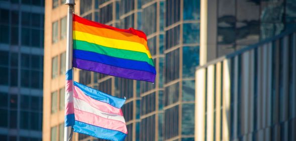 A pride flag is flying above a trans pride flag on a flagpole in an urban setting.