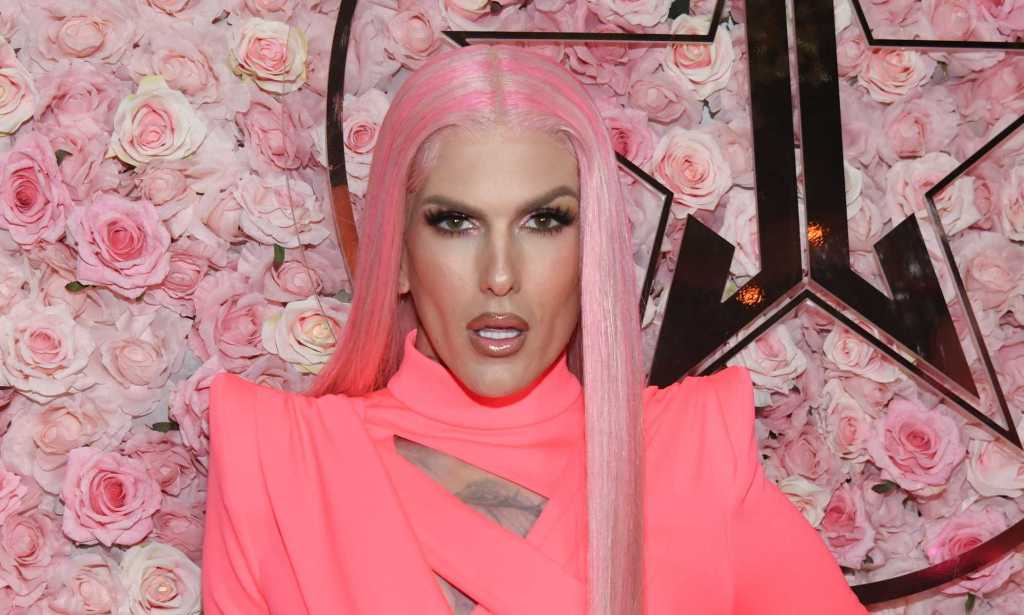 Photo of makeup artist Jeffree Star wearing a pink dress and pink wig standing in front of a background of pink roses