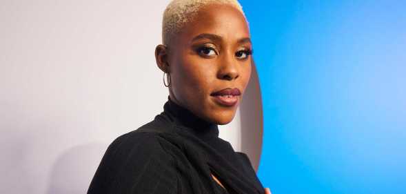 A promo shot of actor Jerrie Johnson wearing a black dress with short white-blonde hair standing in front of a blue and white background