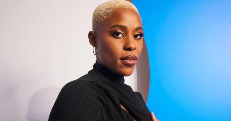 A promo shot of actor Jerrie Johnson wearing a black dress with short white-blonde hair standing in front of a blue and white background