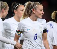 Lionesses captain Leah Williamson stands alongside her football teammates while wearing a white uniform and rainbow OneLove armband