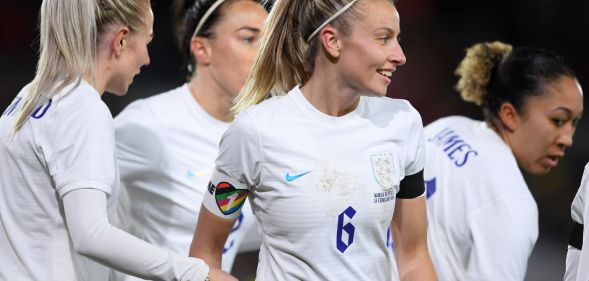 Lionesses captain Leah Williamson stands alongside her football teammates while wearing a white uniform and rainbow OneLove armband