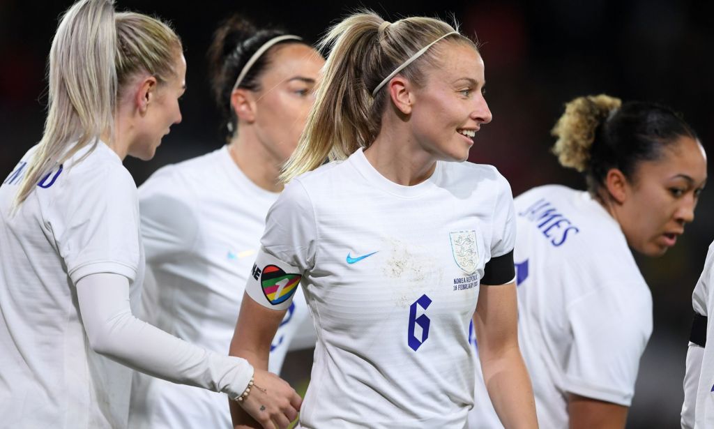 Lionesses captain Leah Williamson stands alongside her football teammates while wearing a white uniform and rainbow OneLove armband