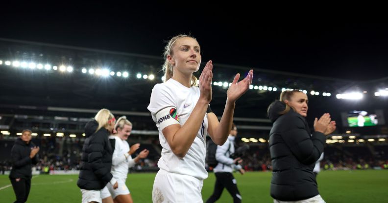 Lionesses captain Leah Williamson claps as she walks off the football field while wearing a white uniform and the rainbow OneLove armband