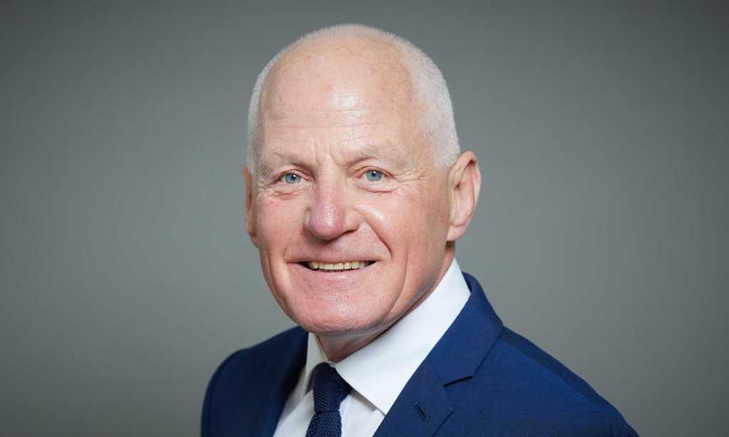 Cashman in a navy suit, white shirt and black tie, smiling against a grey wall