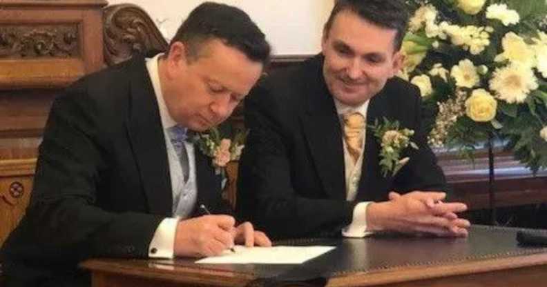 Paul and Alain in suits, sat at a dark wood table, signing their marriage certificate