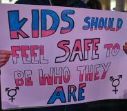 A person holds a sign reading "kids should feel safe to be who they are" in the colour of the trans pride flag