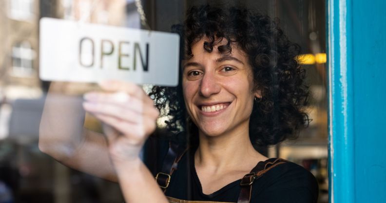 A woman with curly hair is turning over an "open" sign behind a glass door.