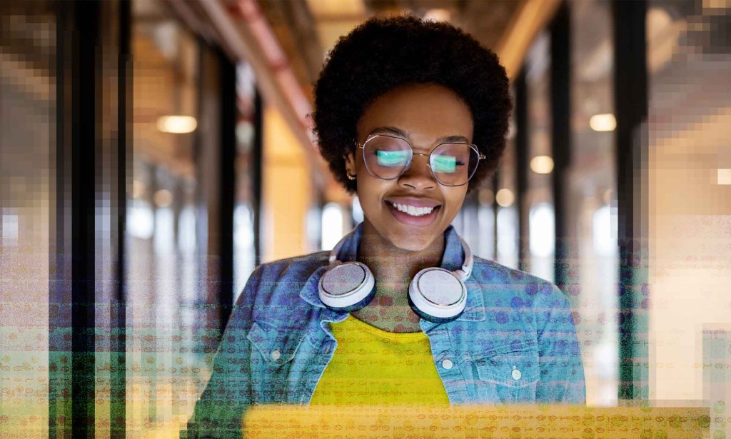 A woman is looking down at her laptop. The reflection of the screen is in her eyeglasses and she has a set of headphone around their neck.