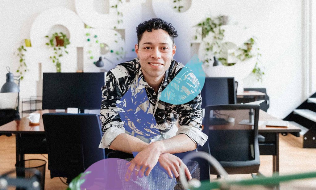 A mand in a floral shirt is leaning on a chair and smiling for the camera. In the background are a computers and plants.