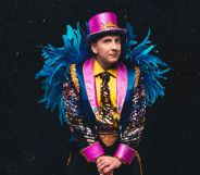 Joe Lycett in a vibrant pink, yellow and blue suit with blue feathers on the arms and a pink top hat.