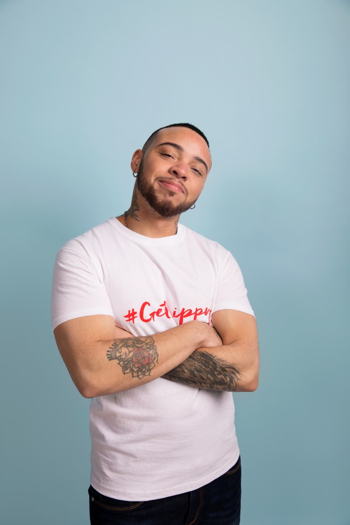 Kenny Ethan Jones is wearing a white t shirt with his arms folded. He has visible tattoos and is standing in front of a blue background.