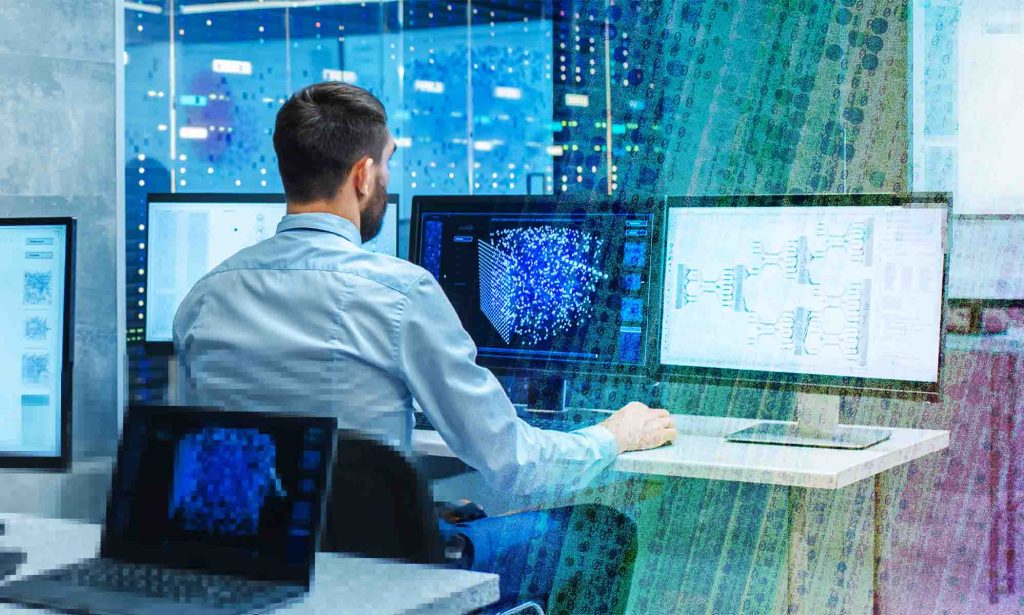 A man in a blue shirt is sitting in front of computer monitors. 