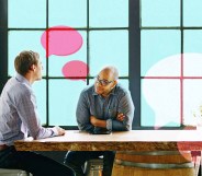 Two men are speaking at a table. One man is wearing classes and looking at the other man. There are illustrations of speech bubbles.