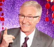 Paul O'Grady in a grey suit and tie, smiling and pointing upwards, against a purple and red background.