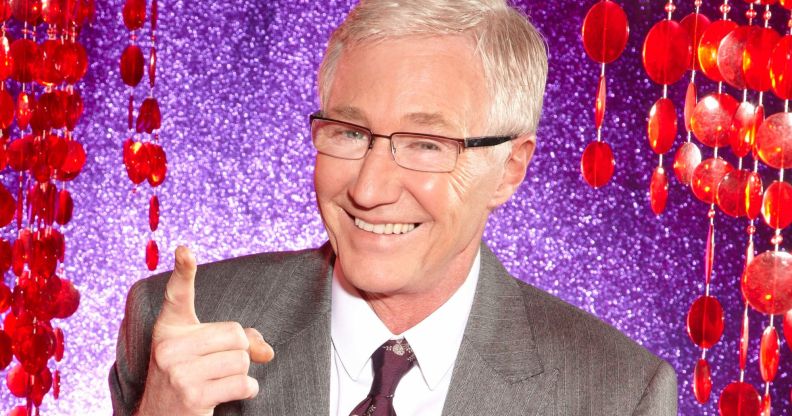 Paul O'Grady in a grey suit and tie, smiling and pointing upwards, against a purple and red background.