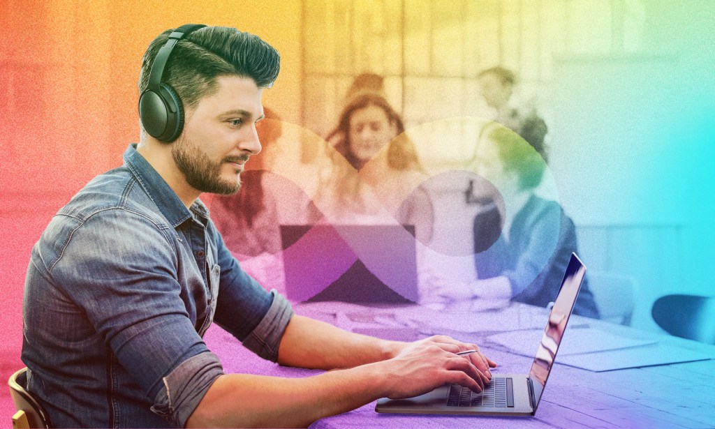 A man wearing a denim shirt is wearing headphones while typing at a computer. There are other people having conversation behind him.