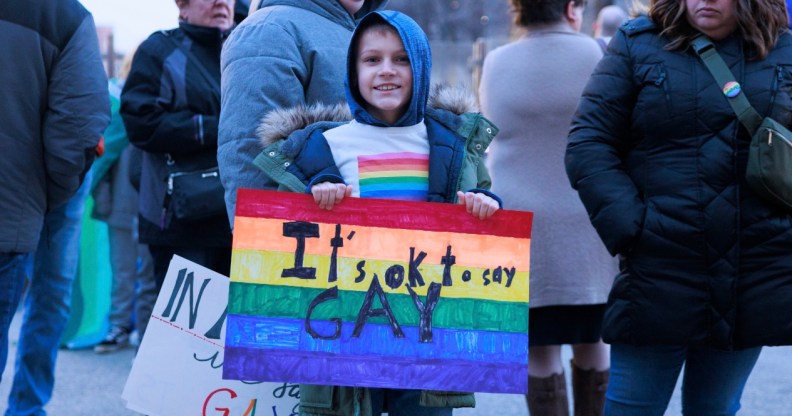 A child in a rainbow T-shirt holds a sign reading "It's okay to say gay"