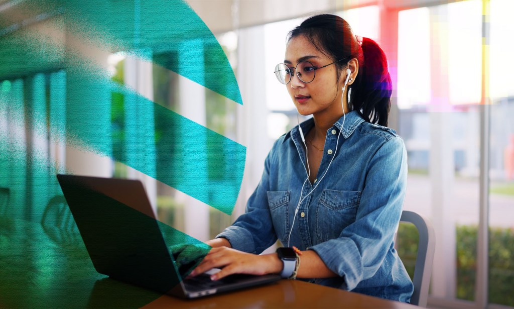 A woman wearing a denim shirt is working on her computer. She has black hair and is wearing headphones. 