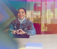 A woman is sitting in a meeting room wearing a denim jacket. There is an inclusive pride flag behind them.