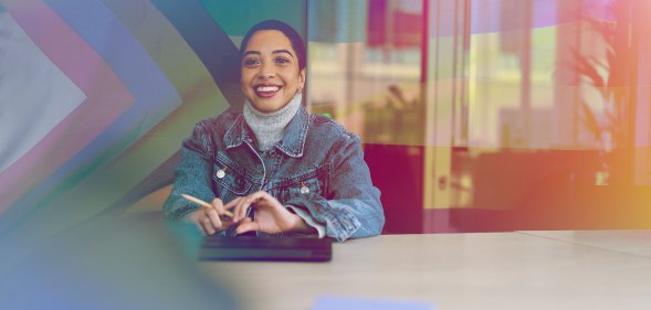 A woman is sitting in a meeting room wearing a denim jacket. There is an inclusive pride flag behind them.