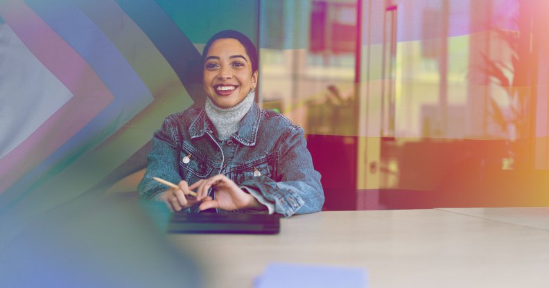 A woman is sitting in a meeting room wearing a denim jacket. There is an inclusive pride flag behind them.