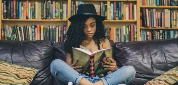 A woman wearing a black hat, blue jeans and black trainers reads a book on a black sofa against a backdrop of books.