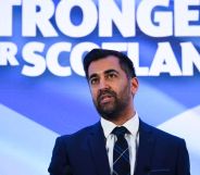 Newly-appointed SNP leader Humza Yousaf wears a suit and tie as he stands in front of a sign in the colours of the Scottish flag