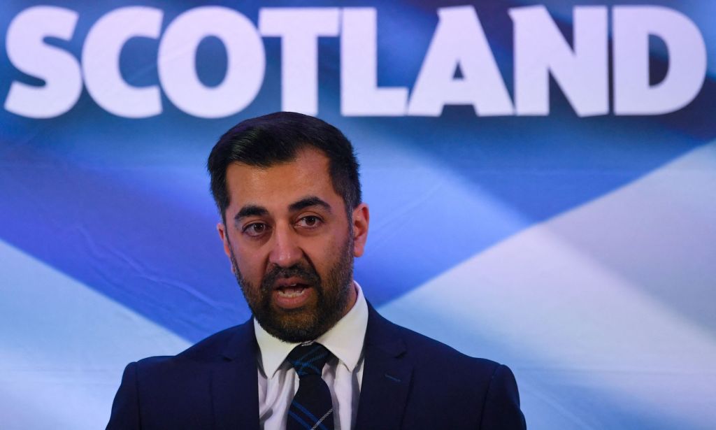 Newly-appointed SNP leader Humza Yousaf wears a suit and tie as he stands in front of a sign in the colours of the Scottish flag