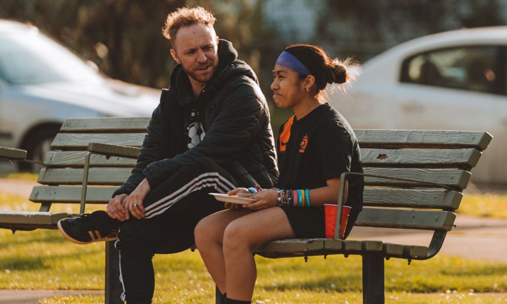 Openly trans coach Kaig Lightner wears a black outfit as he sits on a bench next to another person wearing sports clothings