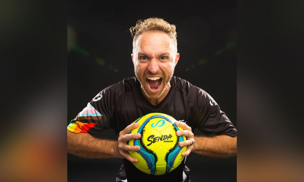 Openly trans coach Kaig Lightner wears a black football jersey with a progressive pride flag on one shoulder as he holds a football in his hands