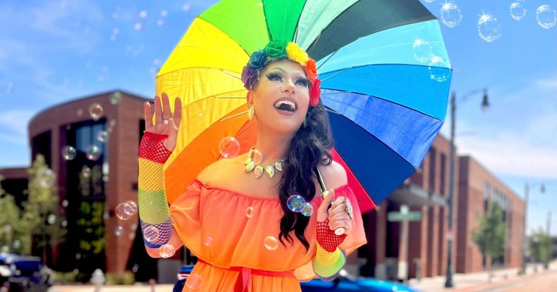 Tennessee drag queen Bella DuBalle, who has been fighting anti-LGBTQ+ bills and the state's drag ban, smiles while holding a rainbow umbrella. She's wearing an orange dress with rainbow accessories