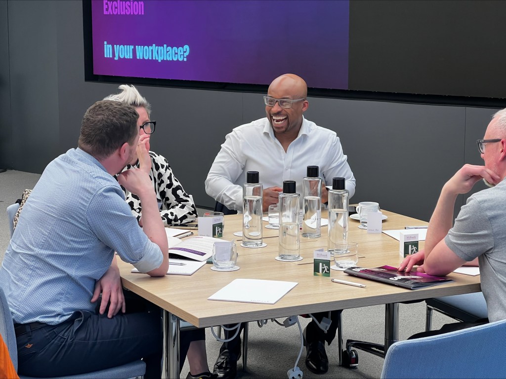 A Black man in a white shirt laughs at a table while colleagues look on. 