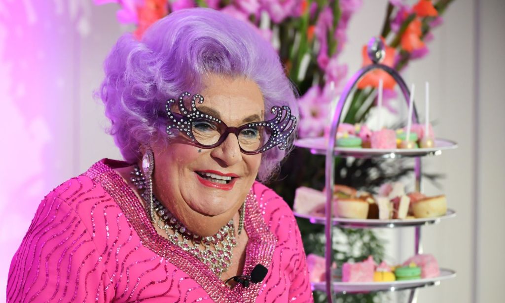 The late Barry Humphries dressed as his drag alter-ego Dame Edna Everage, with purple hair, horn-rimmed glasses and a pink dress