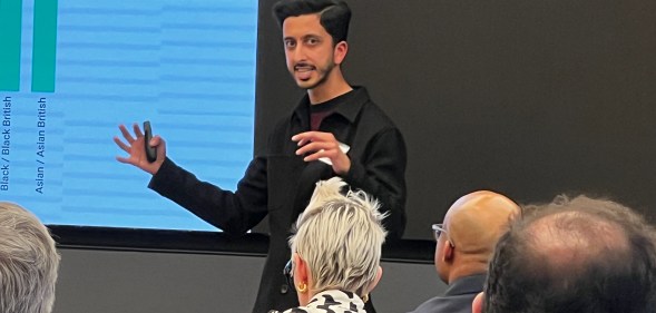 A man in a black shirt is speaking at an event whilst people in the audience look on.