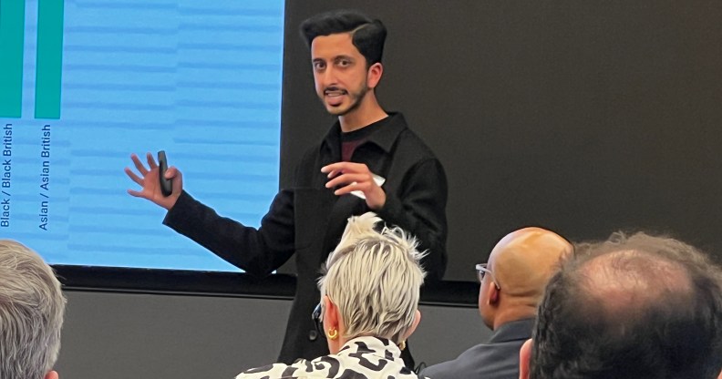 A man in a black shirt is speaking at an event whilst people in the audience look on.